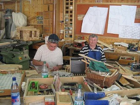 Zwei Bewohner arbeiten in einer Holzwerkstatt, umgeben von Werkbänken und Holzbauteilen; an der Rückwand hängen Baupläne.