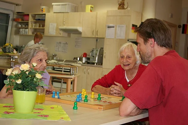 2 Bewohnerinnen spielen 'Mensch ärgere dich nicht' mit einem Mitarbeiter am Küchentisch