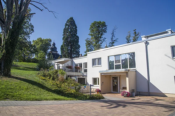 Das 'Haus im Park' der Pflegeeinrichtungen Schloss Meerholz mit gepflastertem Vorplatz und Zugangsweg, dahinter viel Grün