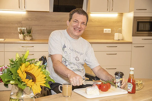 Portrait eines lächelnden Bewohners im Rollstuhl, der sich in der Küche an einem Tisch mit Schneidebrett und Messer eine Mahlzeit zubereitet