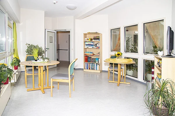 Heller Raum mit Zugang zum Wintergarten. Im Raum sind Bücherregal, Sitzgelegenheiten und ein Fernseher